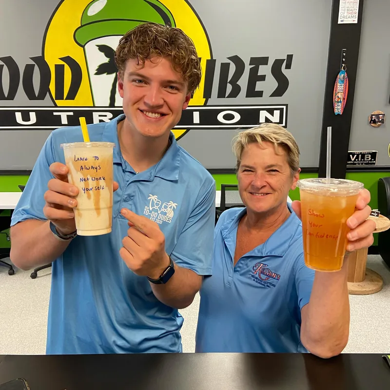 Two smiling customers in blue shirts hold up their drinks in front of the Good Vibes Nutrition wall; one cup reads 'Learn to always not doubt yourself', the other 'Show yourself love'.