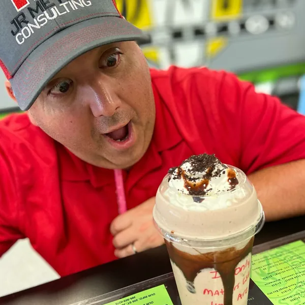 A man in a red shirt and cap grins at a chocolate milkshake topped with whipped cream and cookie crumbles.